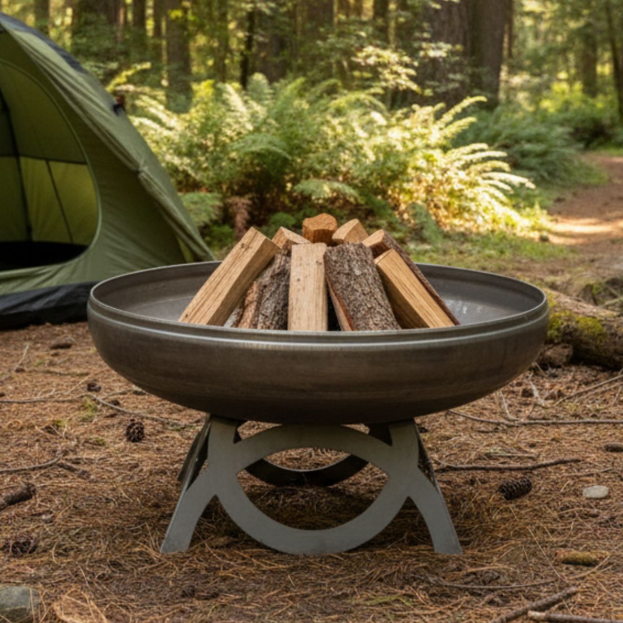 The Liberty Fire Pit - Curved Base - with wood and camping gear in a natural setting.