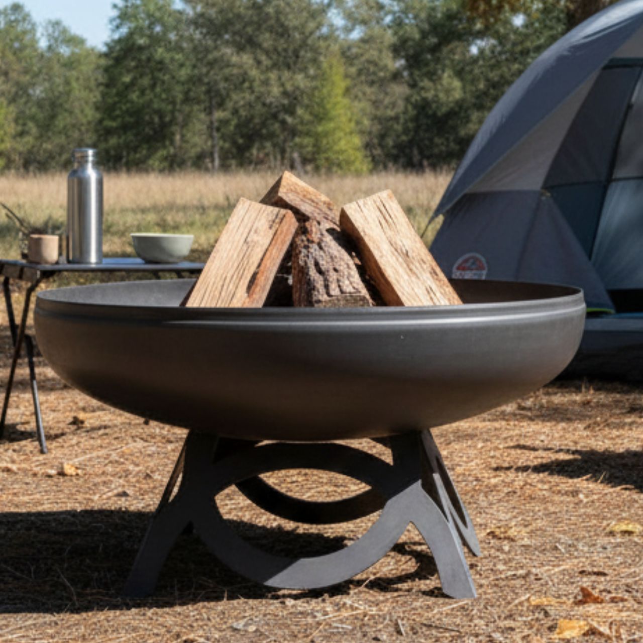 The Liberty Fire Pit - Curved Base - with wood and camping gear in a natural setting.