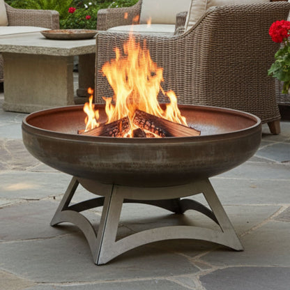 The Liberty wood burning carbon steel fire pit with hollow base, with flames in a patio setting next to chairs and plants.