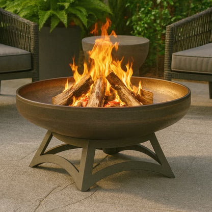 The Liberty wood burning carbon steel fire pit with hollow base, with flames in a patio setting next to chairs and plants.