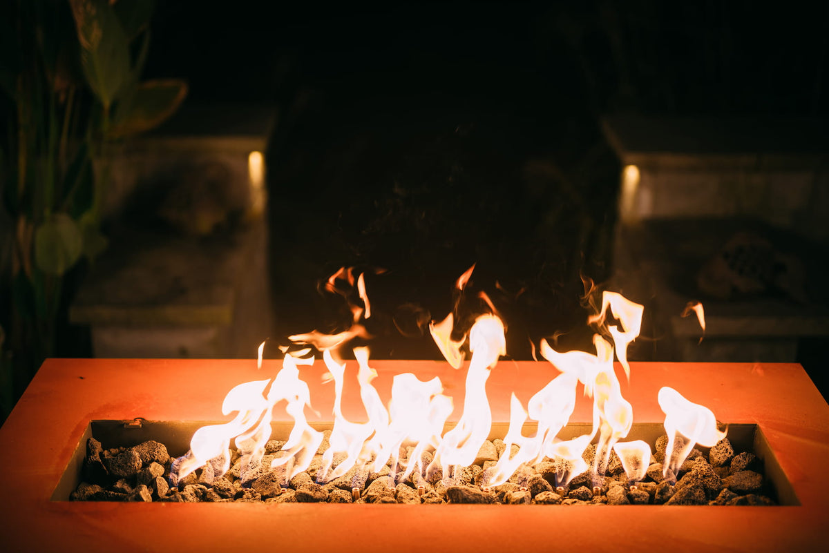 Linear fire pit table with flames and rocks against a dark background.