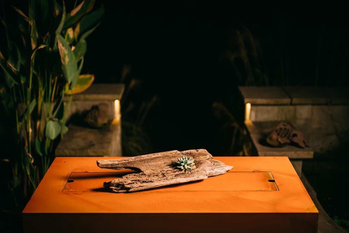 Rectangular Linear fire pit table with driftwood and a small succulent resting on its steel lid, glowing in warm patio lighting.