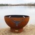 Long Horn 36″ Carbon Steel Fire Pit with weathered patina next to a body of water. 