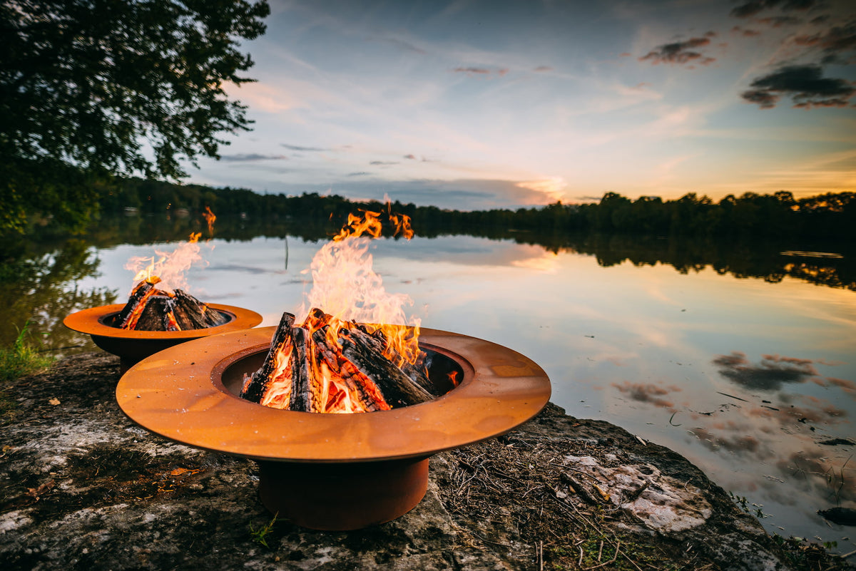 Magnum 54" Steel Fire Pit in foreground with Saturn behind, both burning wood at sunset, by a lake.