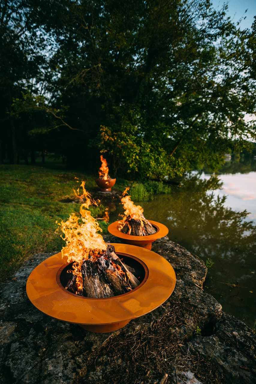 Three fire pits with flames lit up outdoors near a body of water and trees.  Magnum 54" carbon steel fire pit in the foreground with Saturn directly behind.