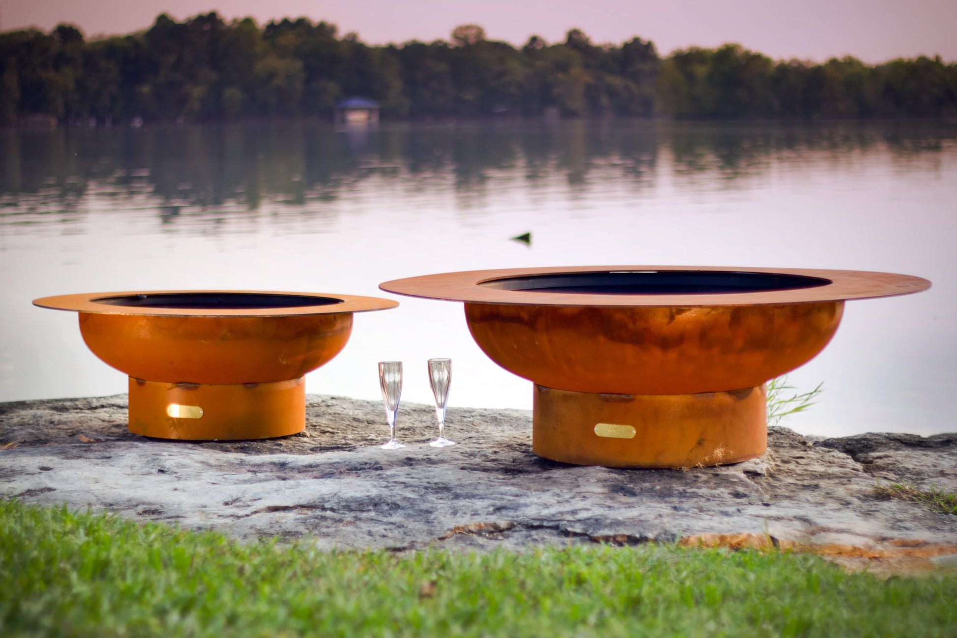 Saturn 41" steel fire pit on left next to Magnum 54" steel fire pit on right with lake in background.