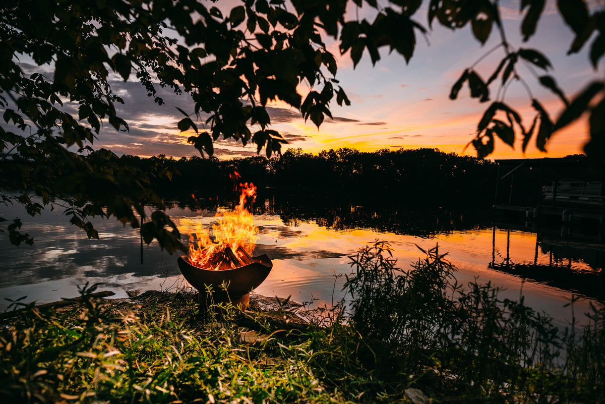 Manta Ray 36" carbon steel fire pit with blazing firewood by a lake at sunset with trees and water in the background.