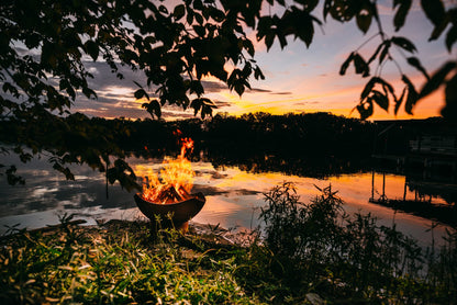 Manta Ray 36" carbon steel fire pit with blazing firewood by a lake at sunset with trees and water in the background.