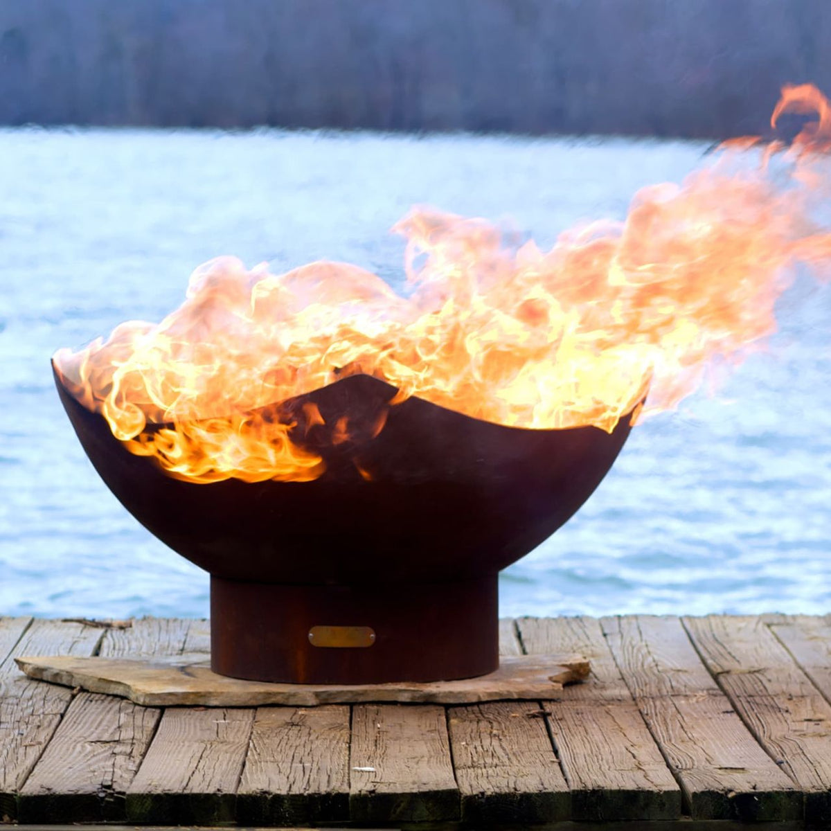 Manta Ray 36-inch carbon steel fire pit with flames, displayed on a wooden dock near a lake.  Made in the USA by Fire Pit Art.