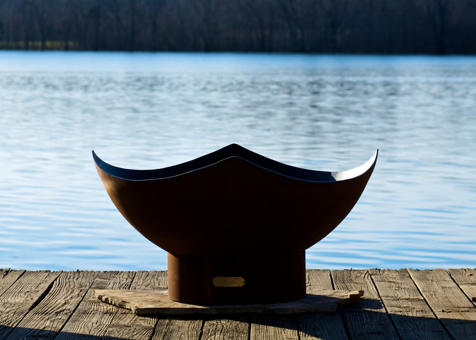 Sideview of unlit Manta Ray 36" carbon steel fire pit in daylight by a lake.