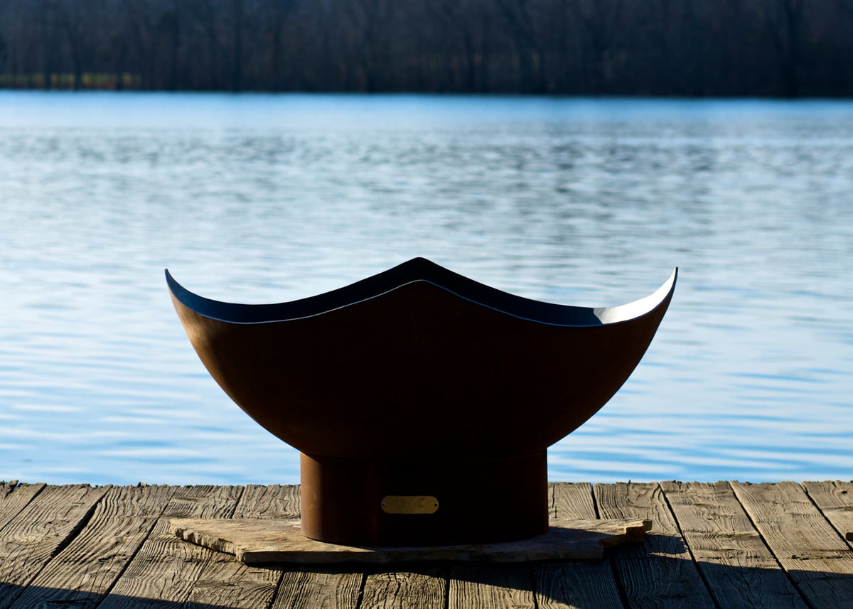 Sideview of unlit Manta Ray 36" carbon steel fire pit in daylight by a lake.