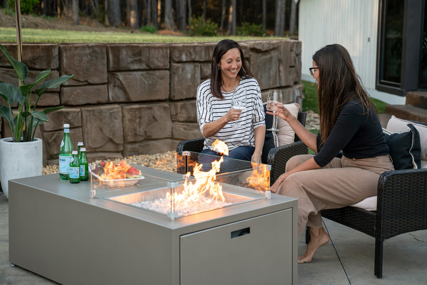 Two female friends toasting with drinks beside The Milan Propane Fire Table - Offset in Champagne on a cozy evening (wind shield sold separately). Shown in a retired finish — see swatches for current Endura options.