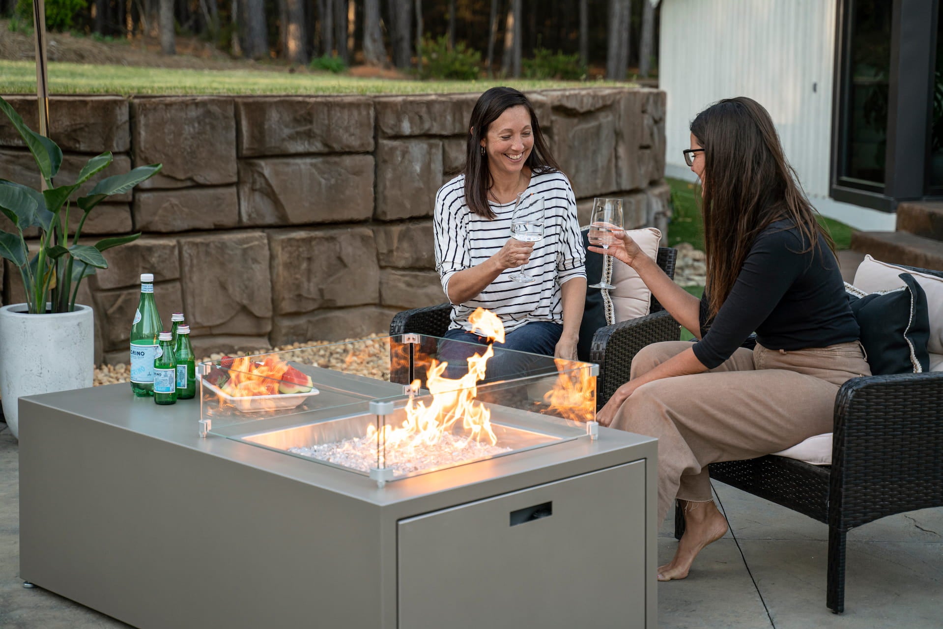 Two female friends toasting with drinks beside The Milan Propane Fire Table - Offset in Champagne on a cozy evening (wind shield sold separately). Shown in a retired finish — see swatches for current Endura options.