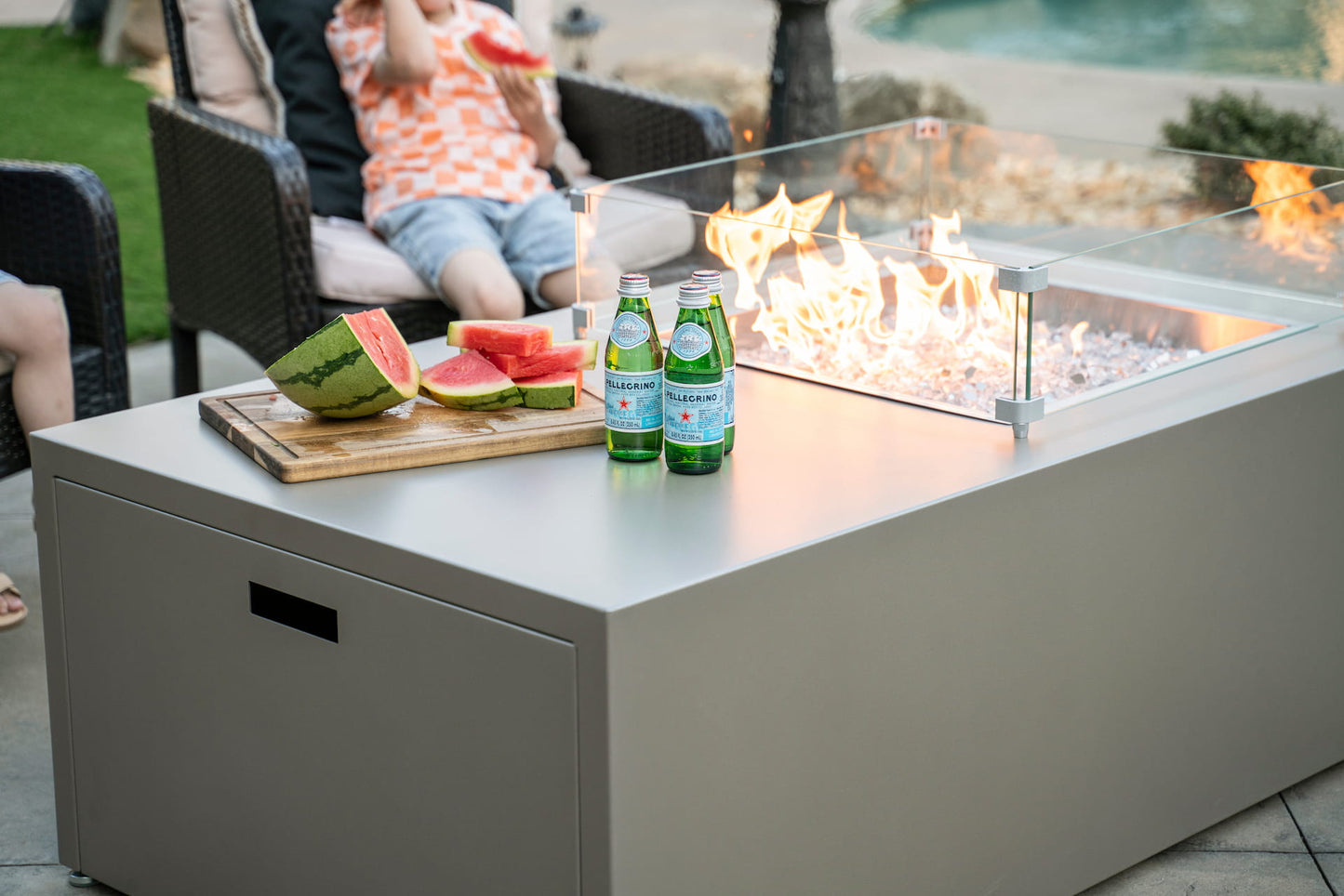 Square wind shield (sold separately) encloses the fire feature of The Milan Propane Fire Table - Offset in Champagne with sparkling drinks and watermelon on the tabletop.  Shown in a retired finish — see swatches for current Endura options.