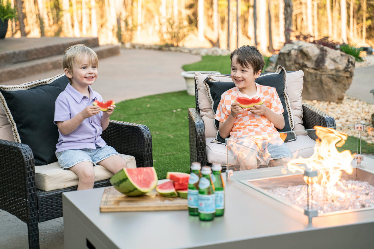 Two smiling kids enjoying watermelon around The Milan Propane Fire Table - Offset on a sunny day (pictured in Champagne, wind shield sold separately). Shown in a retired finish — see swatches for current Endura options.