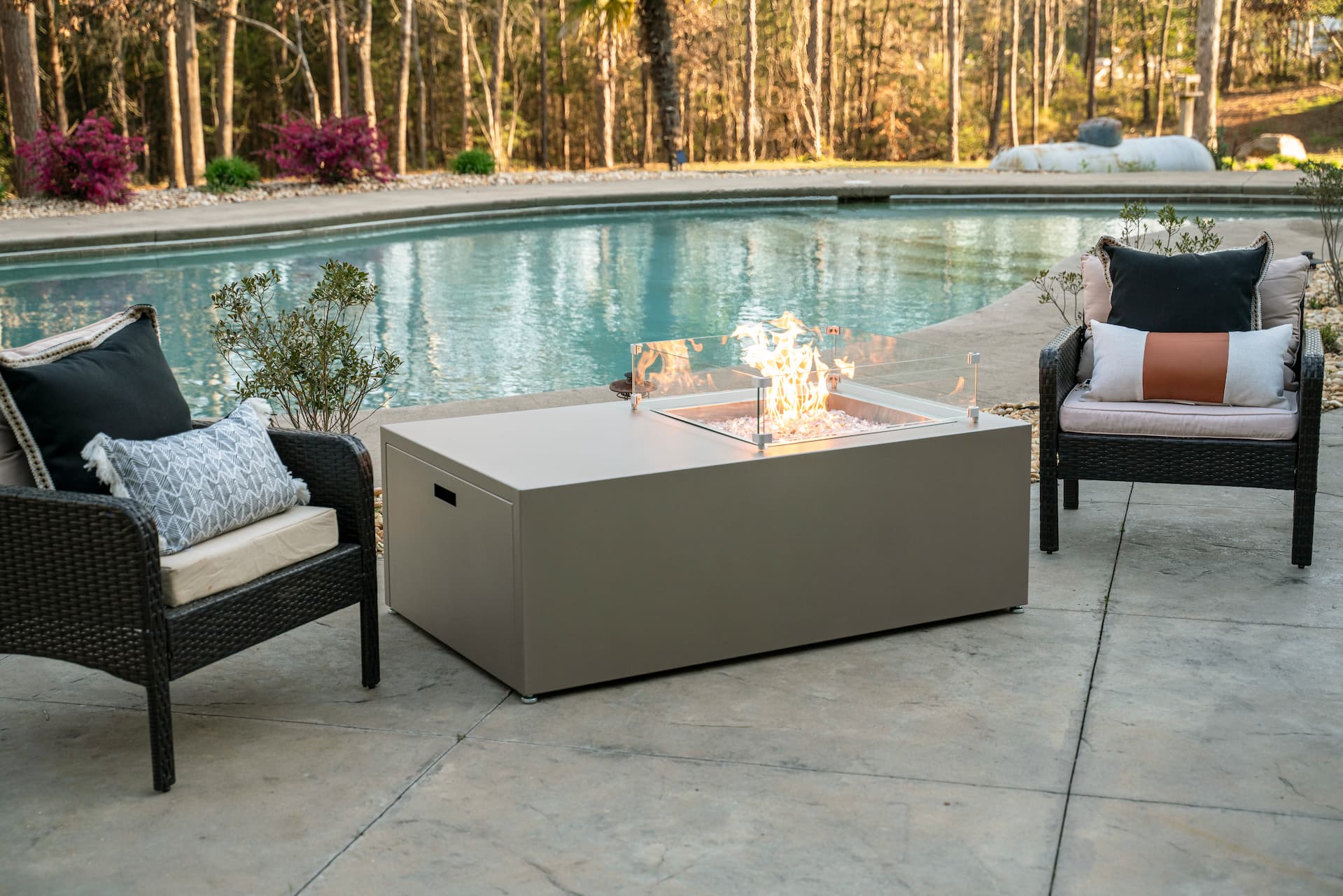The Milan Propane Fire Table - Offset in Champagne on a patio by the pool, surrounded by wicker lounge chairs (wind shield sold separately). Shown in a retired finish — see swatches for current Endura options.