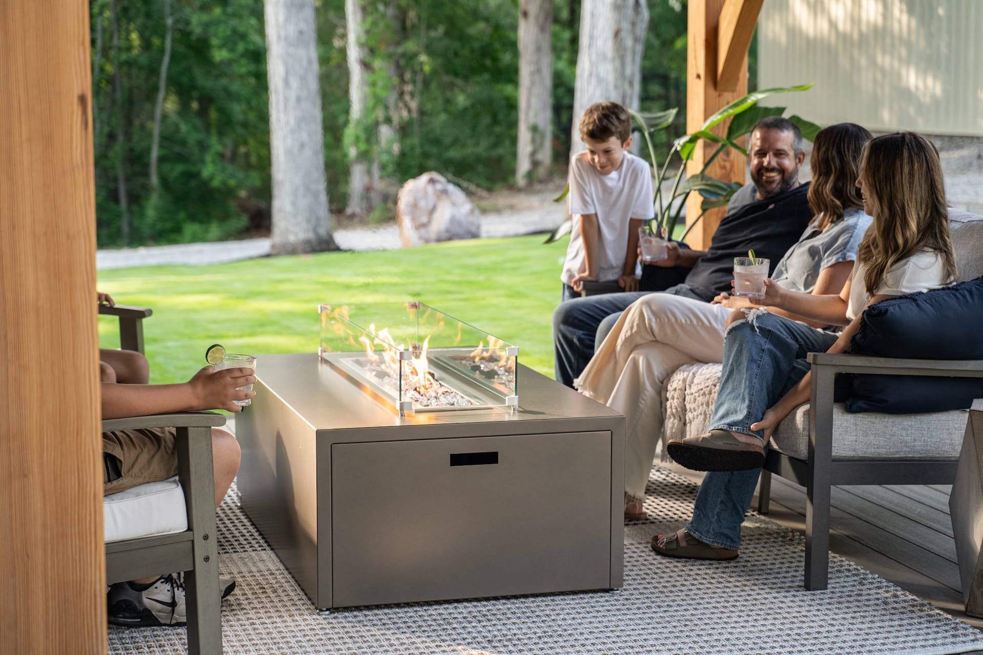 Family enjoying conversation around Milan Linear propane fire table in Endura Concrete finish (wind shield sold separately).
