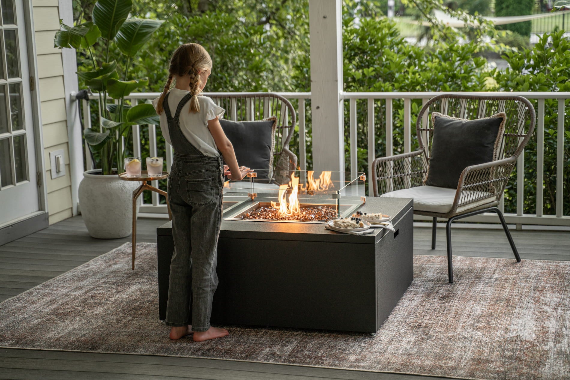 Child standing on a deck next to The Milan Squre fire pit with a view of a house and garden and her snacks nearby.