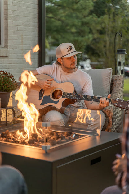 Man playing acoustic guitar next to The Milan Propane Fire Table - Square with warm flames and relaxed vibe (pictured in Graphite, wind guard sold separately).