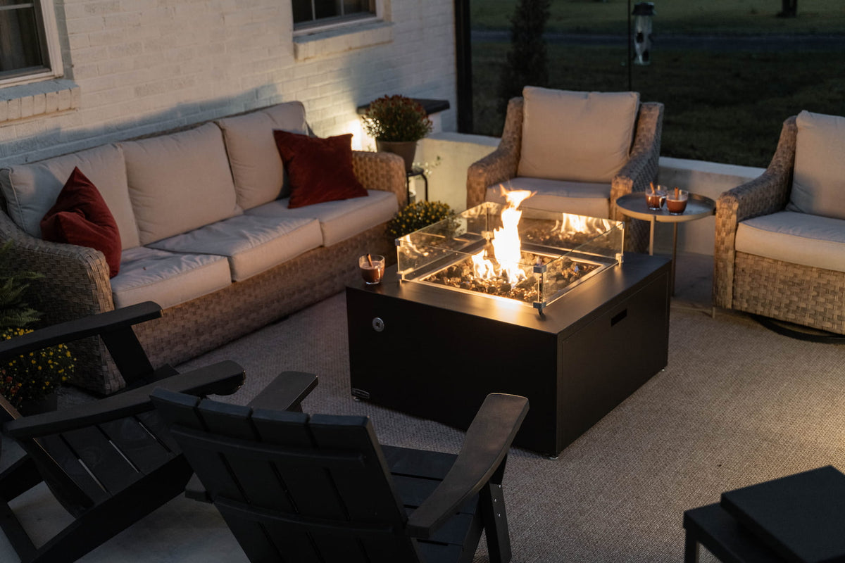 Evening patio setup with The Milan Propane Fire Table - Square lit and surrounded by cozy wicker furniture (pictured in Endura Graphite, wind shield sold separately).

