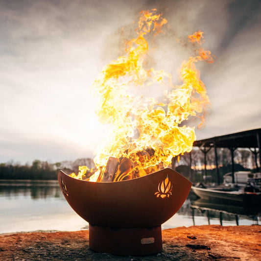 Namaste' 36 inch carbon steel fire pit with blazing wood in front of lakeside dock.