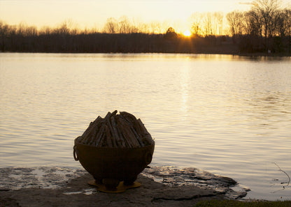 Fire Pit Art's Nepal 41" carbon steel fire pit lakeside at sunset with unlit wood inside.
