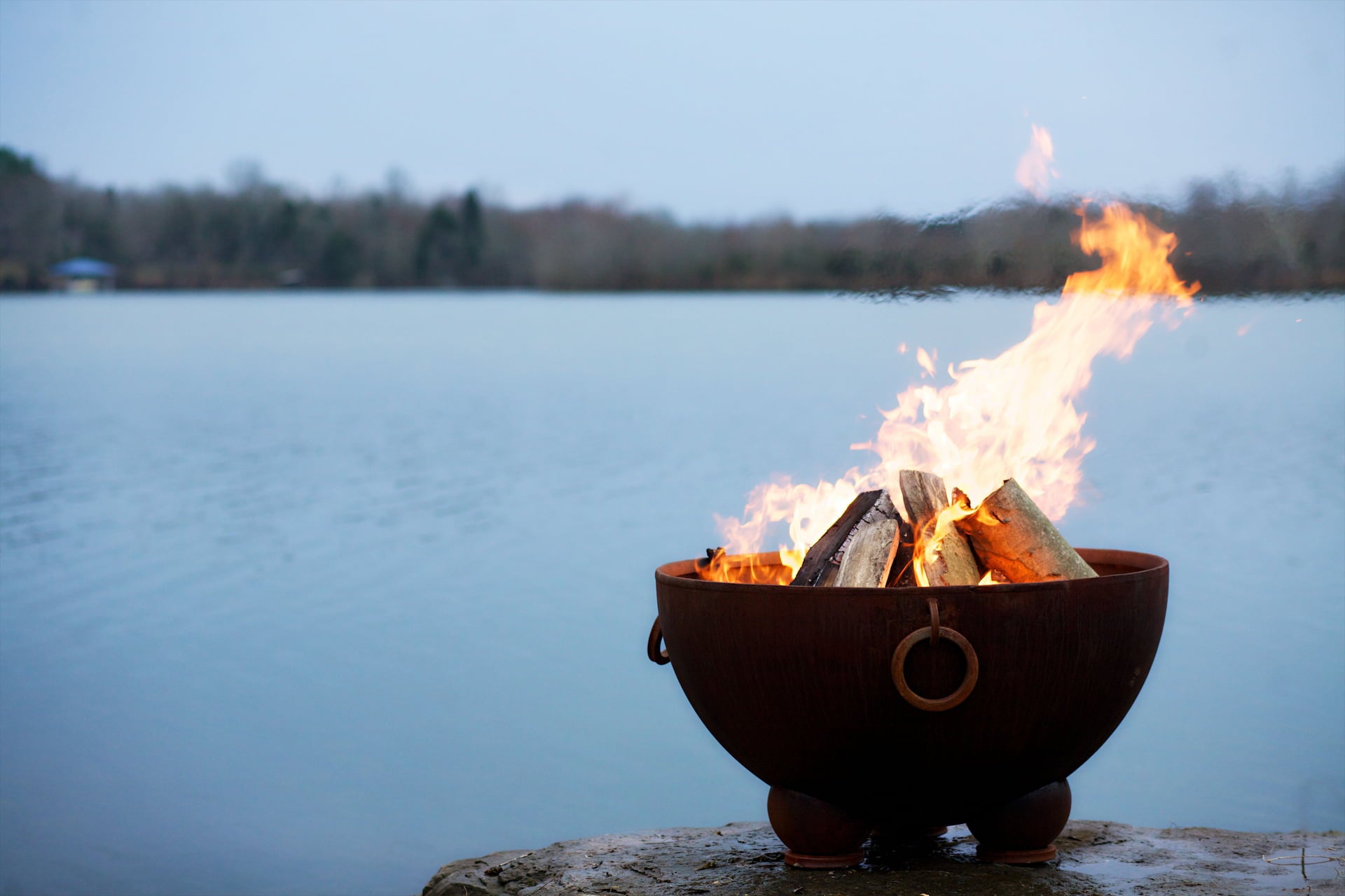 Nepal fire pit filled with burning logs, placed lakeside at dusk ,with calm water in the background. Made in the USA  by Fire Pit Art.