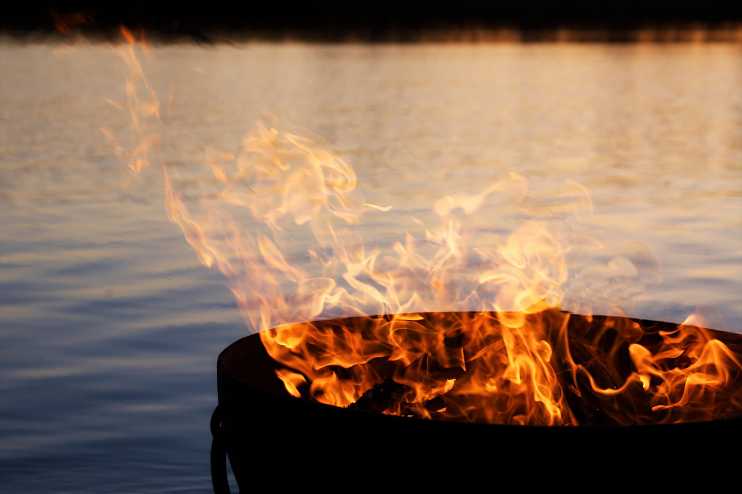 Dramatic flames rising from Nepal fire pit at sunset with water reflecting firelight in the background.