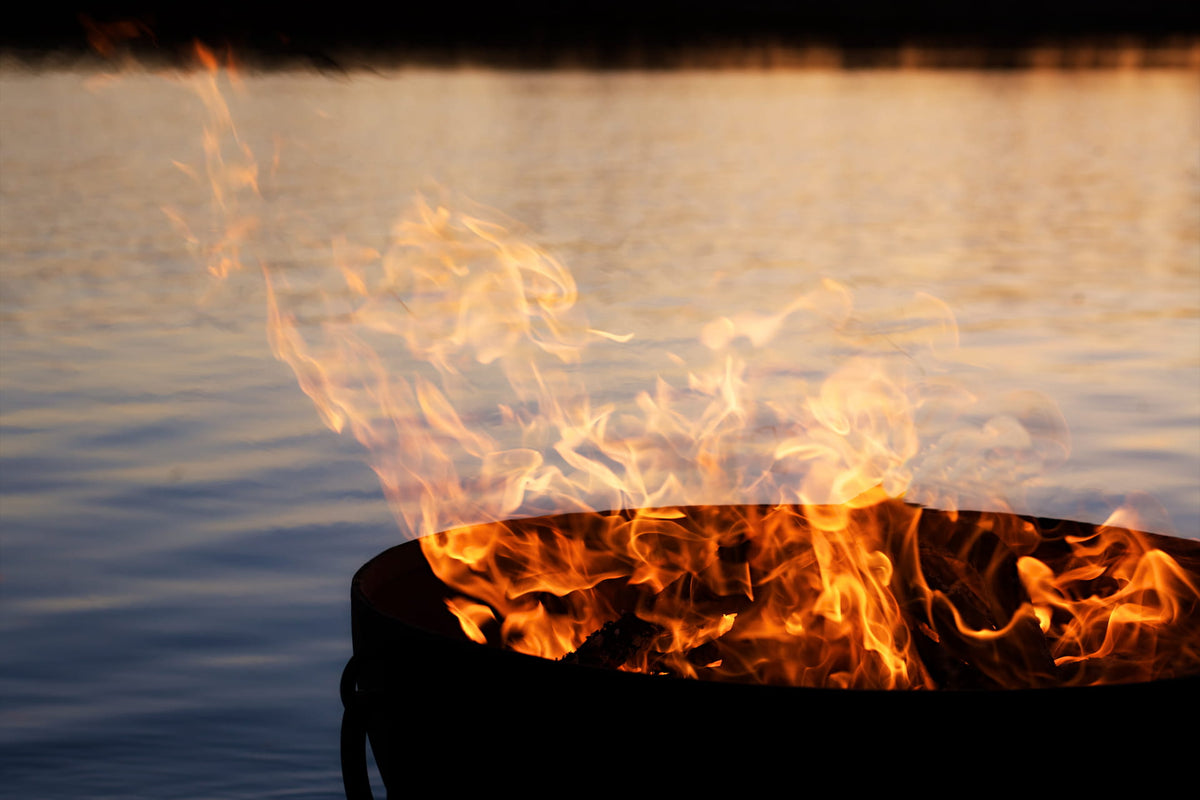 Dramatic flames rising from Nepal fire pit at sunset with water reflecting firelight in the background.