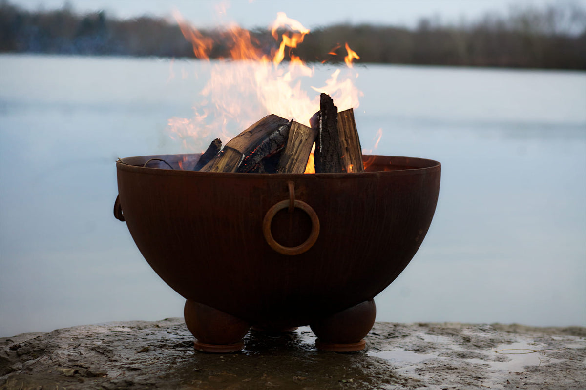 Nepal fire pit in use near a calm lake, with wood fire burning and iron ring detail visible on the bowl.