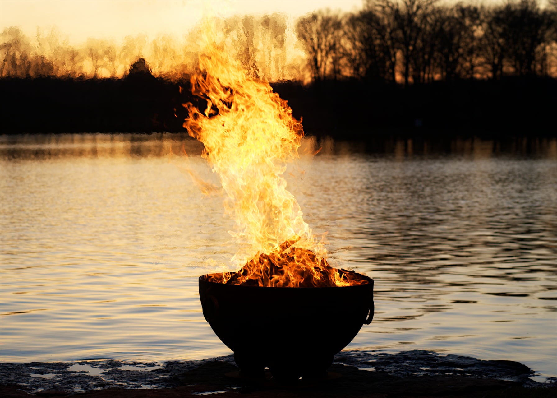 Nepal 41" carbon steel fire pit blazing with tall flames at sunset beside a calm lakeshore. Made in USA by Fire Pit Art.