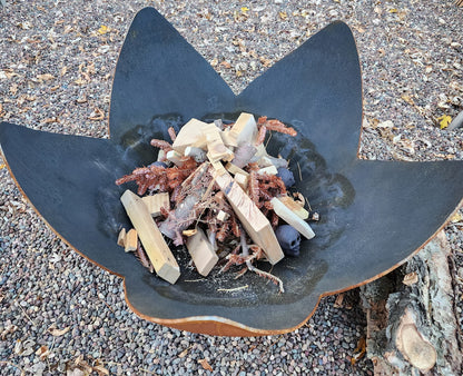 Petal-shaped steel fire pit filled with kindling and wood, ready for lighting in a rocky outdoor setting.
