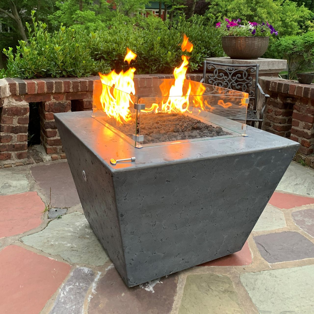 Redan GFRC Concrete fire pit in Rustic Grey with flames on a stone patio with greenery in the background.
