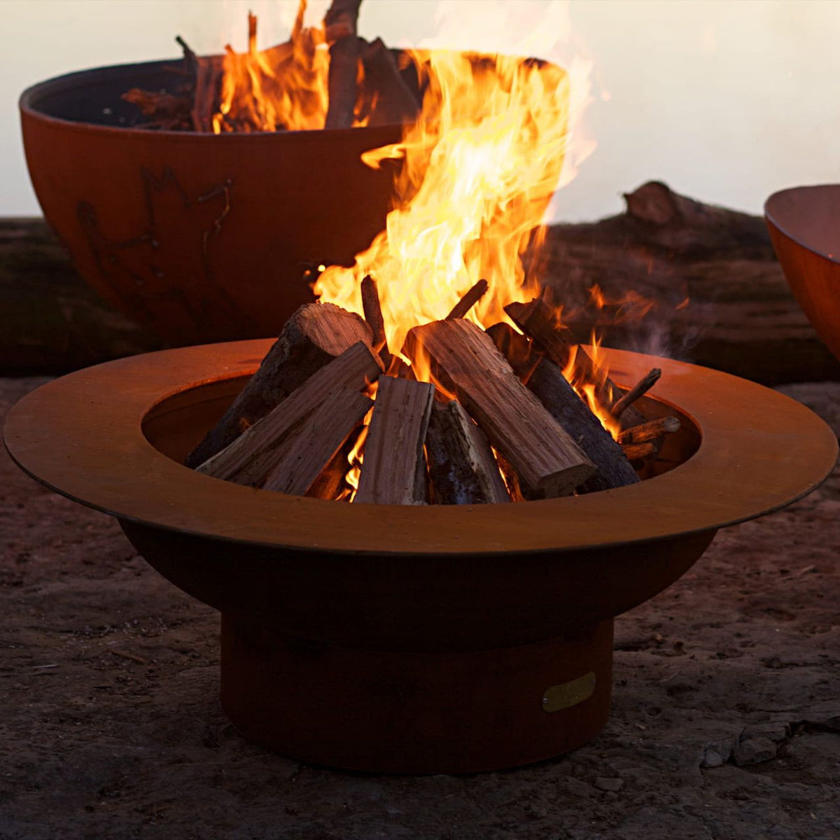 Saturn 41" Steel Fire Pit glowing at sunset, logs ablaze inside the wide steel ring, with Funky Dog fire pit in the background.  Made in USA by Fire Pit Art. 