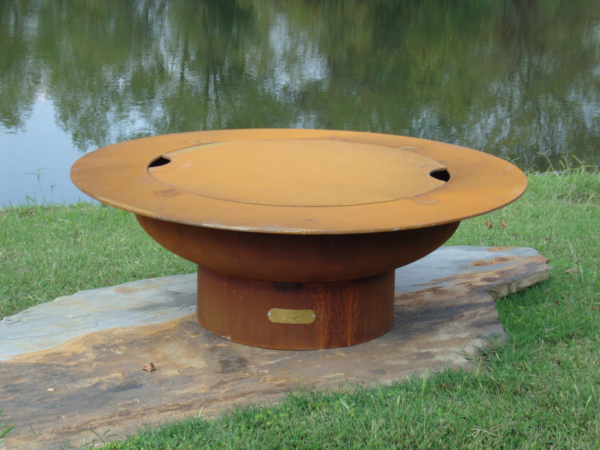 Saturn 41-inch carbon steel fire pit with lid in place, shown on a stone slab surrounded by grass near a calm lakeside.  By Fire Pit Art.