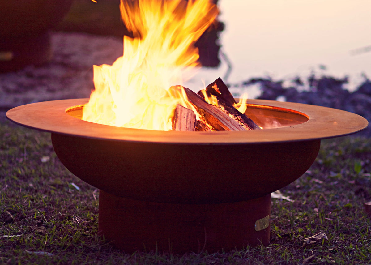 Saturn 41-inch carbon steel fire pit lit with vibrant flames on a grassy lawn at dusk, showcasing the circular rim design and glowing wood fire ambiance.