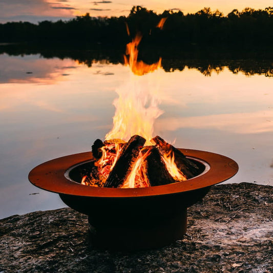 Open wood fire burning in the Saturn 41-inch carbon steel fire pit at sunset by a lake, highlighting the fire bowl’s wide celestial-inspired rim and rustic patina finish.