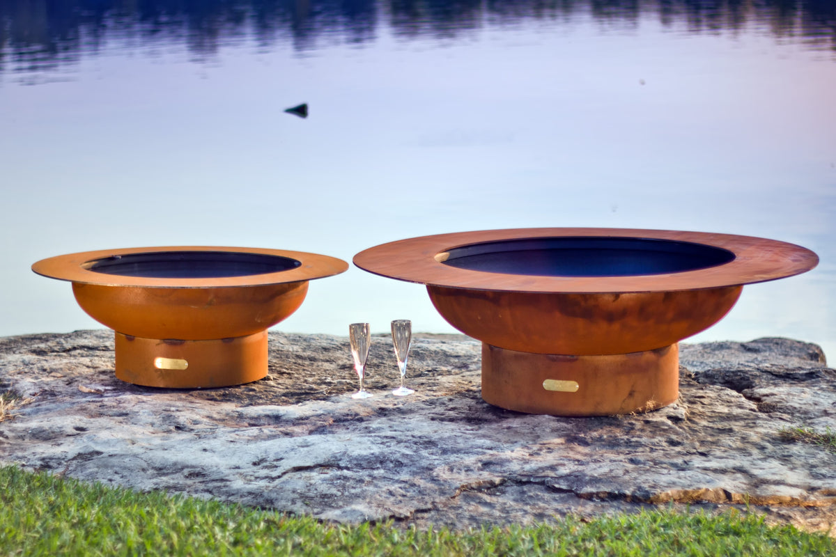 The Saturn and Magnum carbon steel fire pits, side by side, next to a lake.