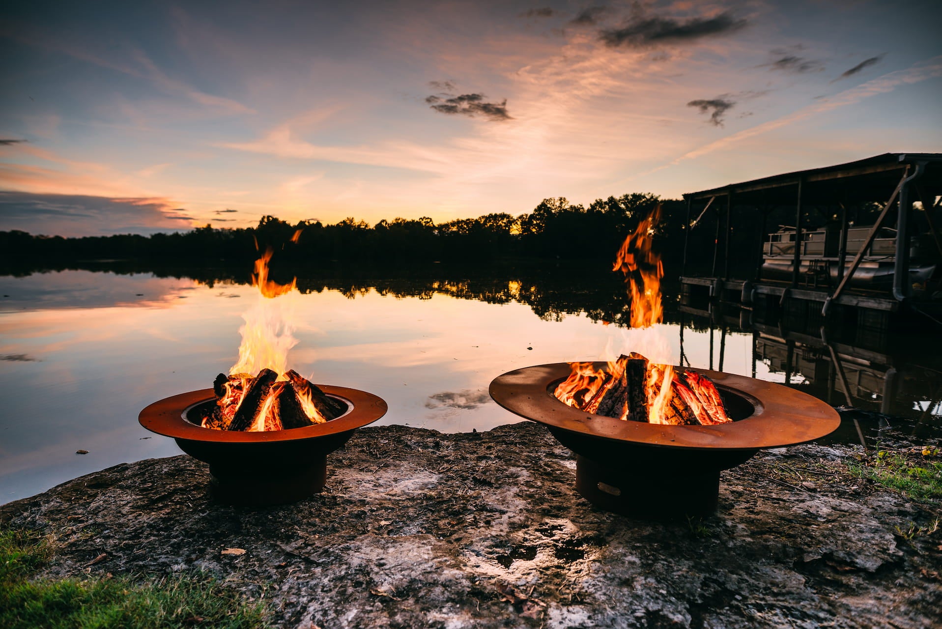 Saturn and Magnum fire pits burning side by side at sunset, showing size and flame style comparison in a lakeside setting.