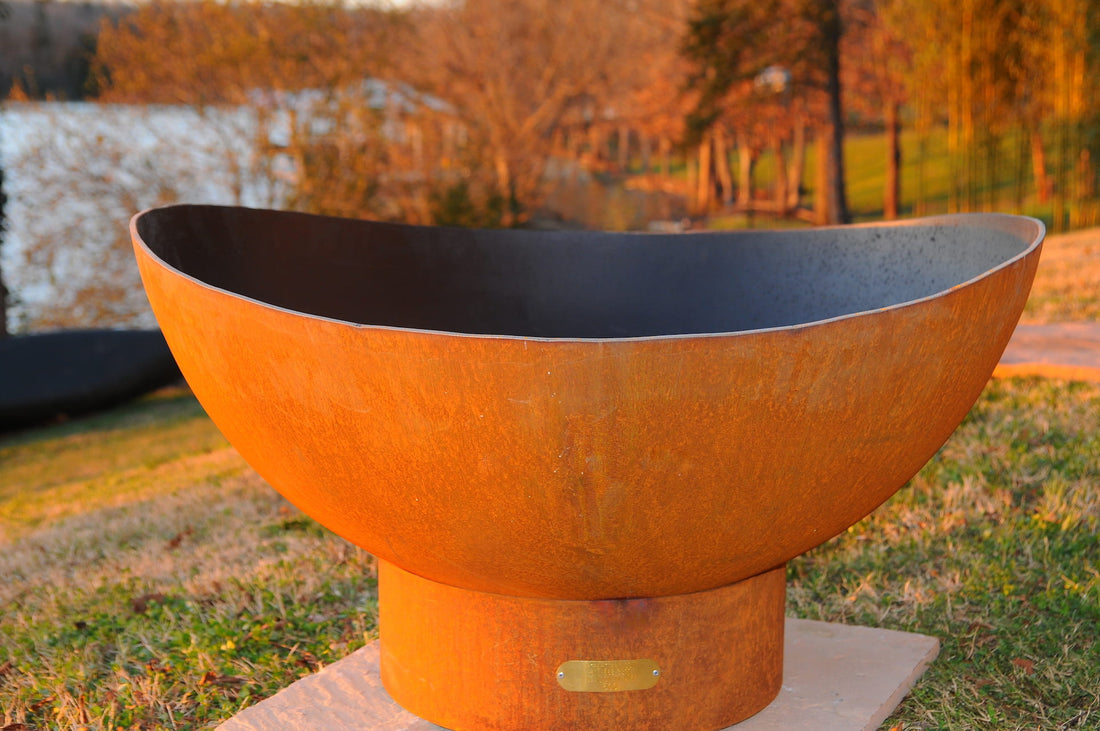 Scallop 36-inch fire pit with rust-toned patina finish and curved rim, displayed on stone slab, with a lake in the distance.