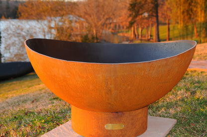 Scallop 36-inch fire pit with rust-toned patina finish and curved rim, displayed on stone slab, with a lake in the distance.