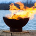 Scallop 36-inch carbon steel fire pit roaring with flames on a dock, set against a peaceful lake with trees in the background.  Made in USA by Fire Pit Art.