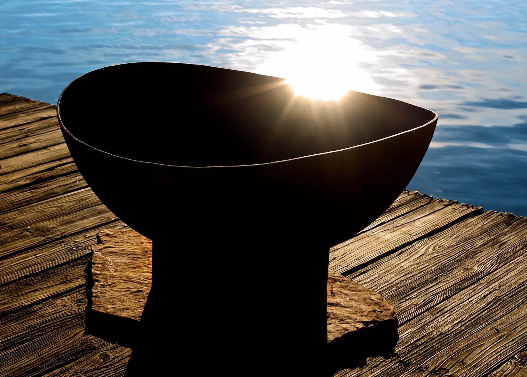 Scallop fire pit silhouetted by the setting sun on a dock with water in the background.