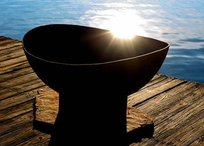 Scallop fire pit silhouetted by the setting sun on a dock with water in the background.