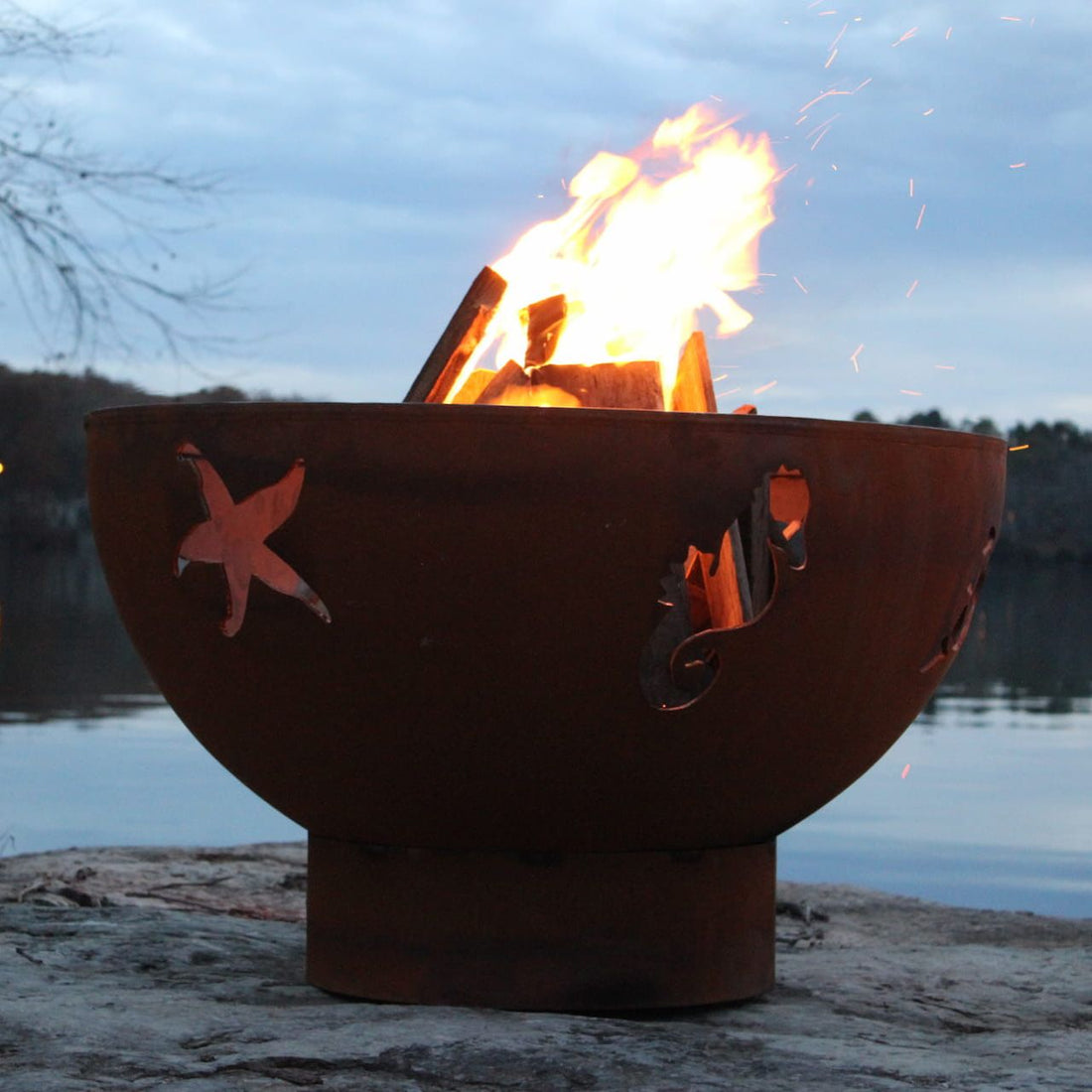 Sea Creatures 36-inch carbon steel fire pit with ocean cutouts glowing by the lakeside at dusk.  Made in USA by Fire Pit Art.