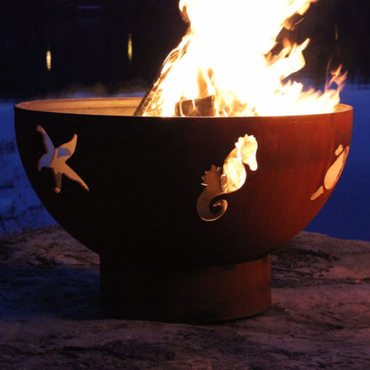 Lit Sea Creatures 36-inch carbon steel fire pit showing starfish and sea horse cutouts with lake in background during evening light.