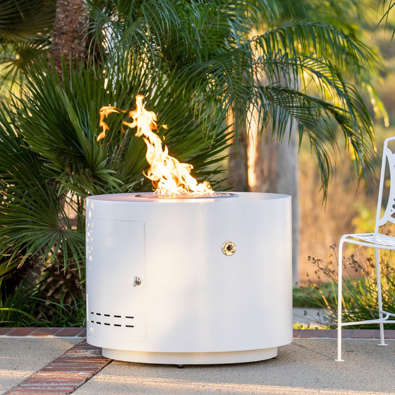 The lit Beverly round steel fire pit in white powder coat on a patio with palm tree in the background.