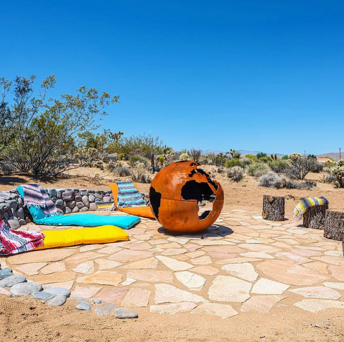Third Rock 36" carbon steel fire pit displayed in a colorful desert lounge with low seating and bright pillows.