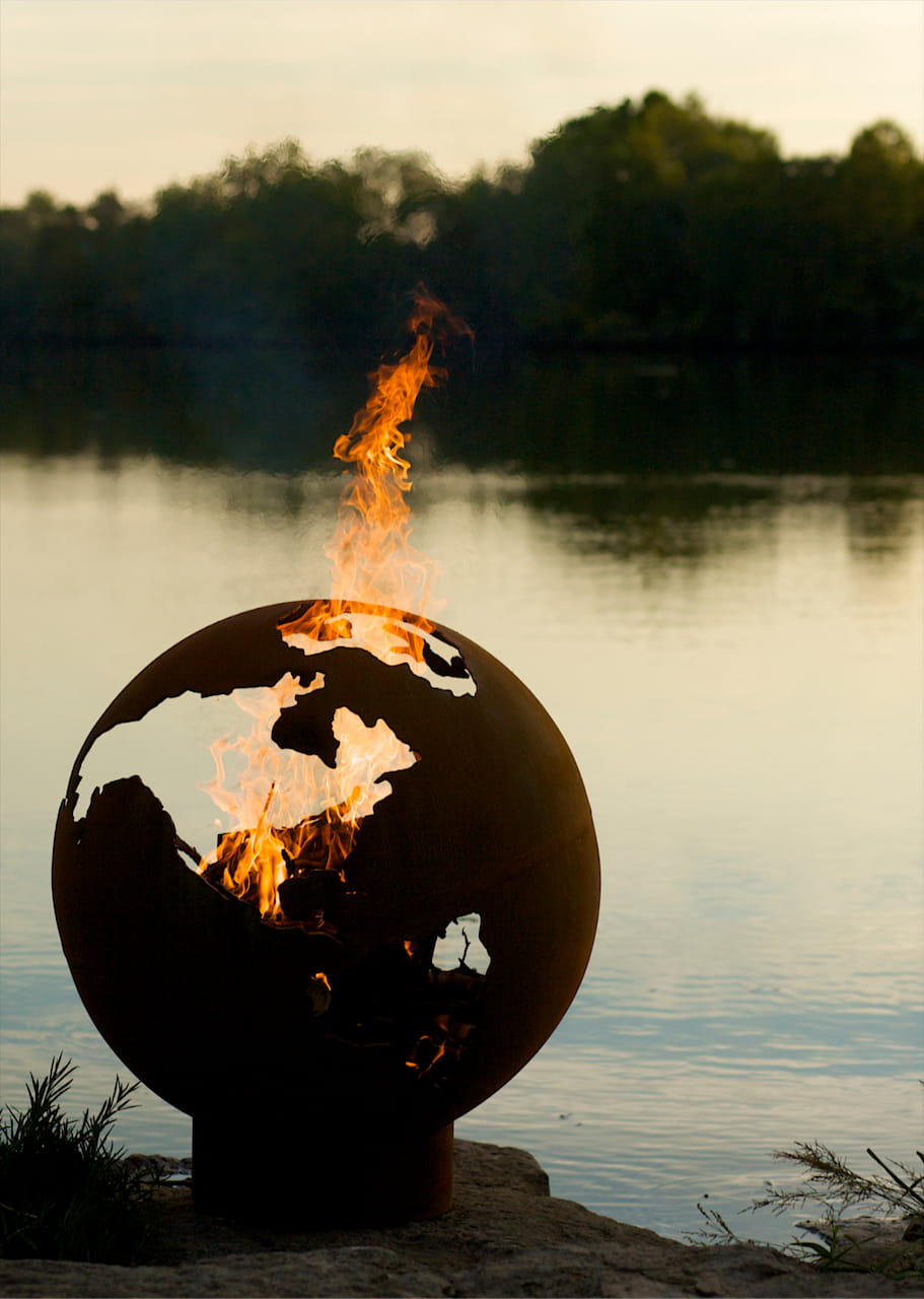 Fire Pit Art’s Third Rock 36-inch steel fire pit by a lake, flames visible through North America cutout.