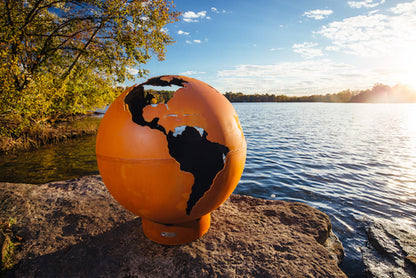 View of the spherical Third Rock 36" carbon steel fire pit on a sunny day next to a lake.